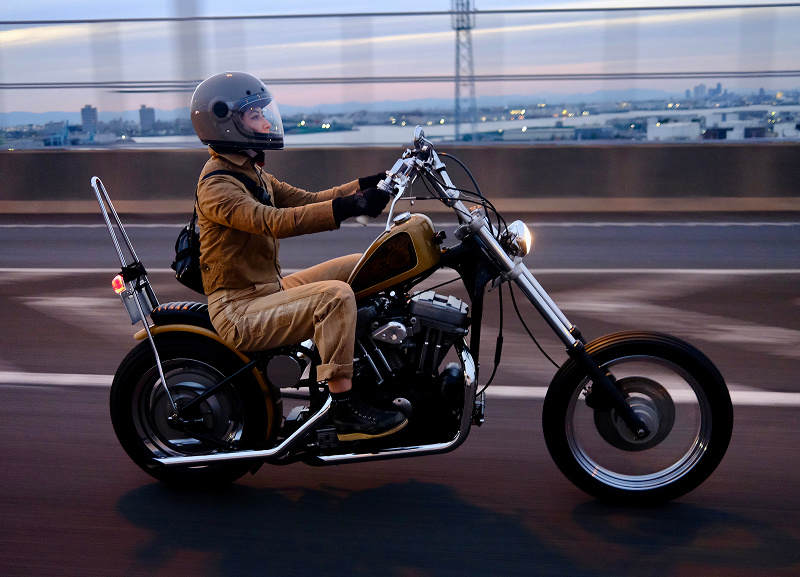 Rider cruising on a low-slung custom chopper along a roadway with a blurred guardrail and water in background