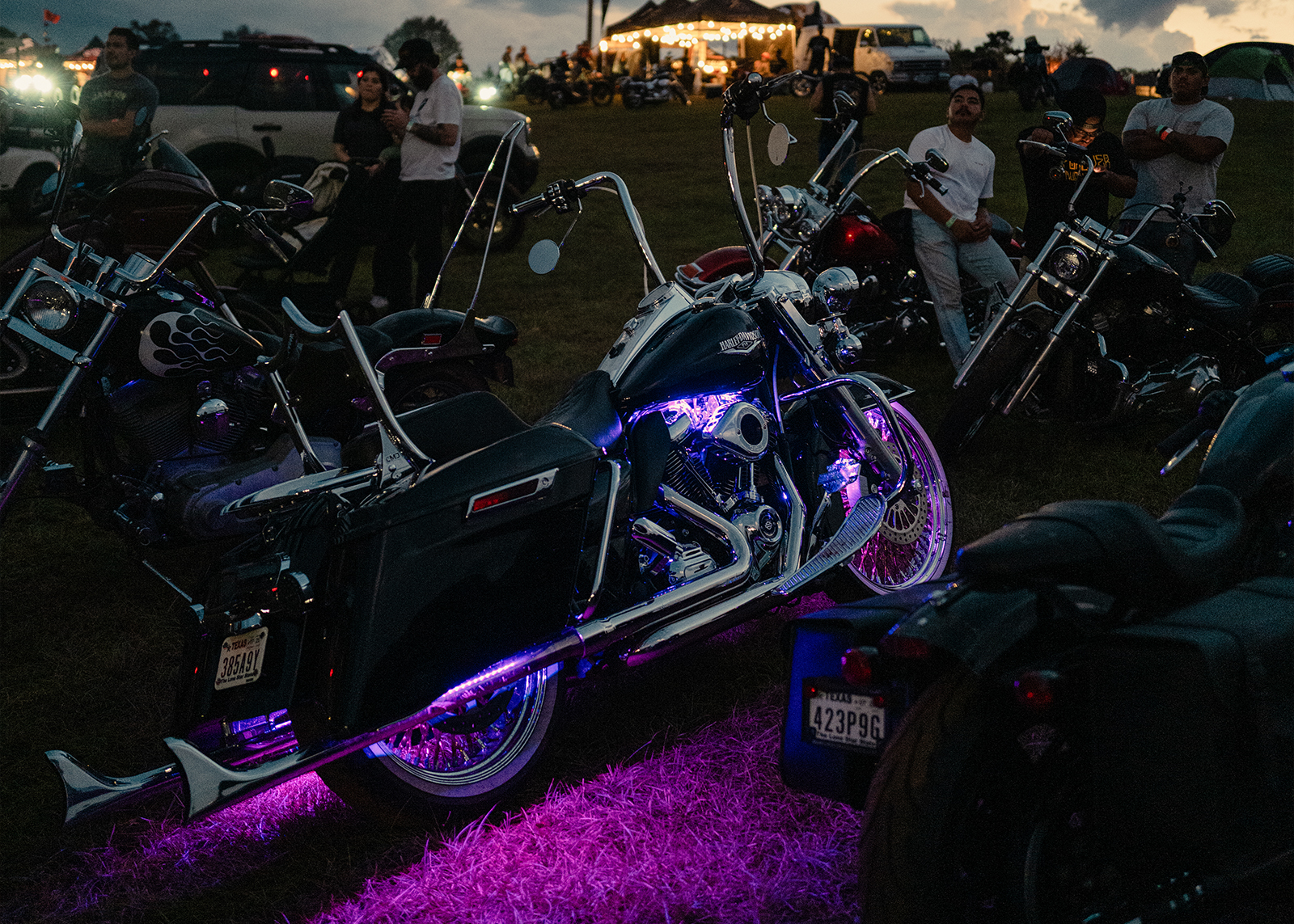A Harley-Davidson glows with purple LED lights at dusk, surrounded by bikes and onlookers at the Born Free Texas rally.