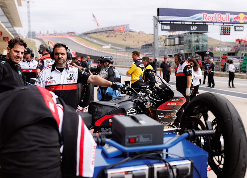 Motorcycle and crew gathered on pit lane with equipment carts as the track and grandstands rise behind