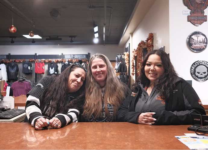 Three employees leaning on a counter inside a Harley‑Davidson retail store, with apparel racks and wall decor behind them