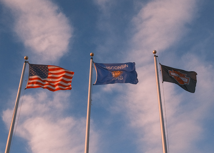 Three flags flying on tall poles—the United States flag, Wisconsin state flag, and Harley‑Davidson flag against a blue sky