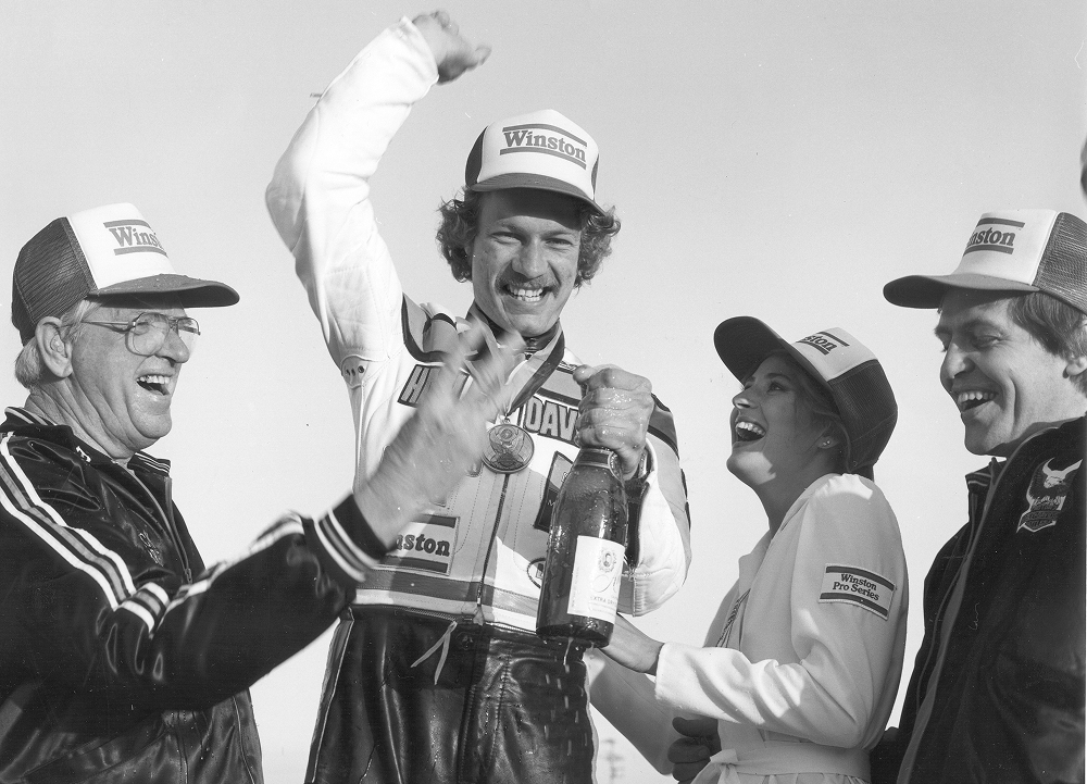 Jay Springsteen celebrates on a podium with a medal, trophy bottle, and supporters