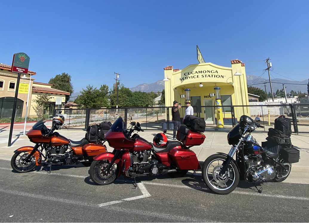 Three motorcycles parked outside a yellow historic service station with mountains visible in the distance