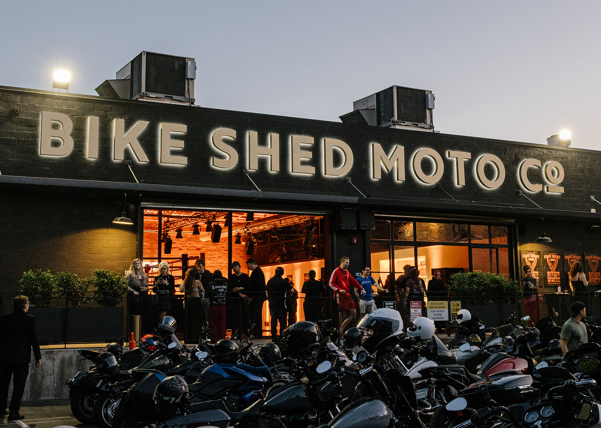 Crowd gathers outside Bike Shed Moto Co, with motorcycles lined up in front under evening lights.