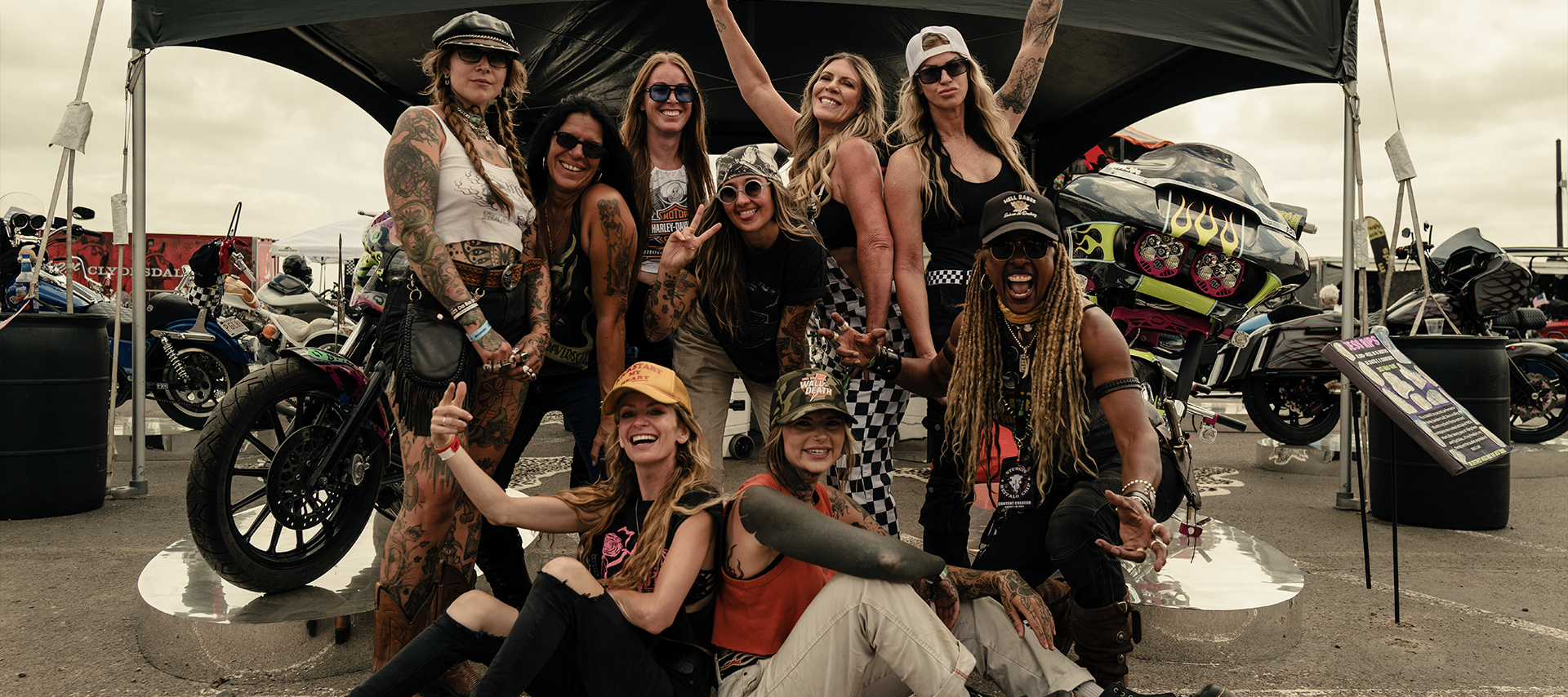 Group of women bikers pose together at a motorcycle event with custom bikes in the background.