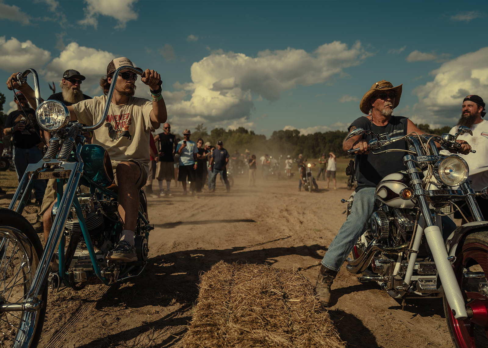 Two riders on vintage choppers line up for a dirt race as a crowd gathers under the bright afternoon sky.