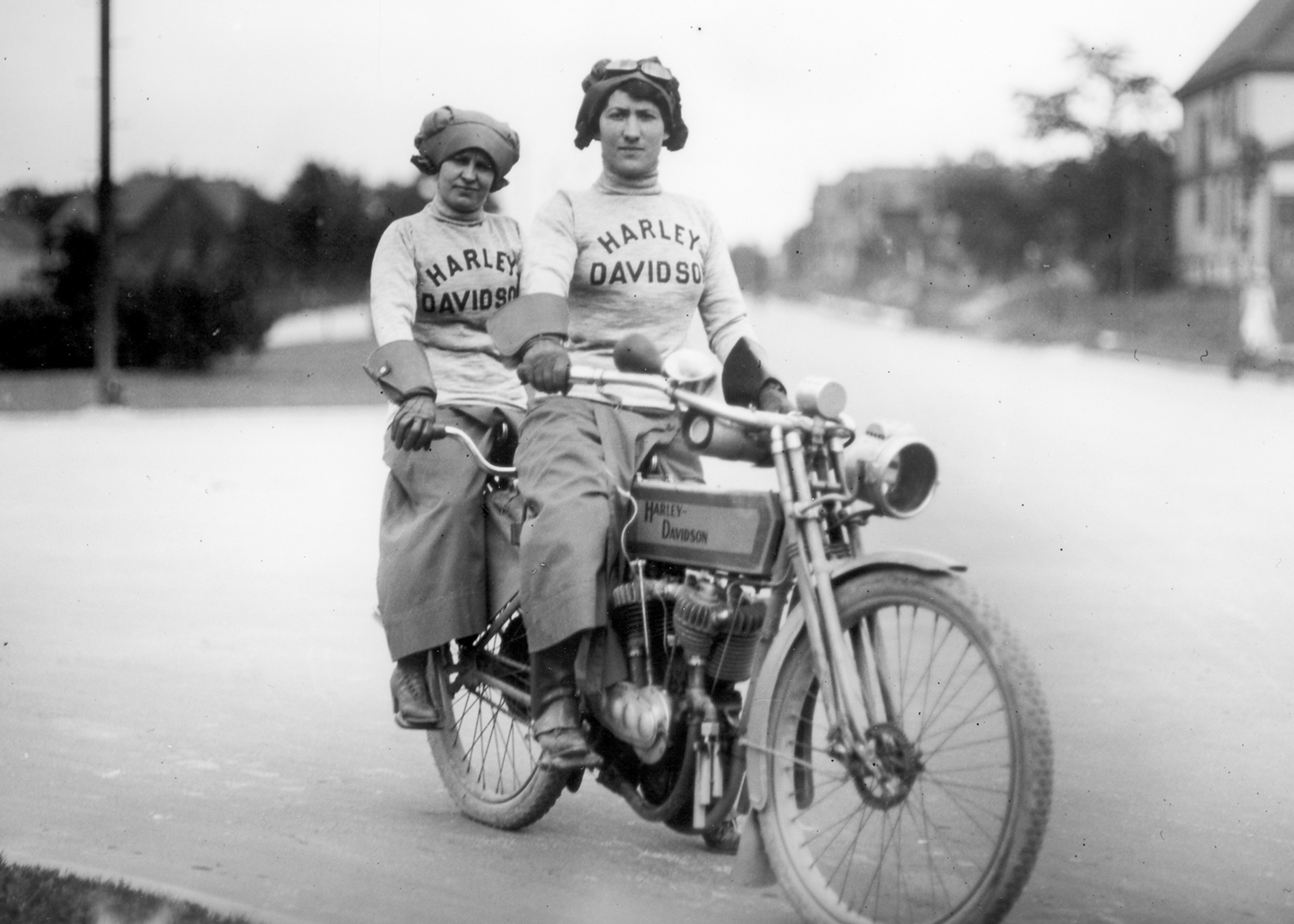 Vintage black-and-white photo of two women wearing Harley-Davidson sweaters ride a vintage motorcycle together on a paved street.