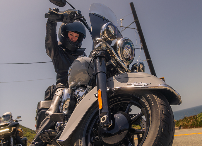 Looking up at rider seated on a cruiser with hand on handlebar parked along a roadside under clear sky