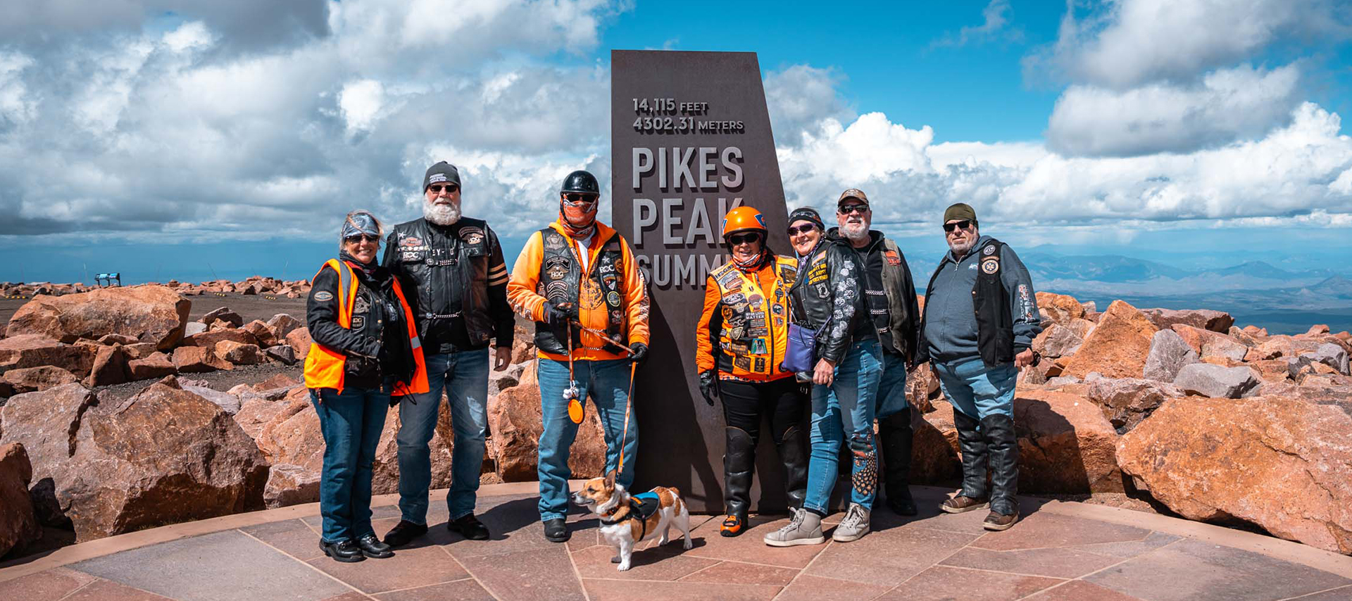 A group of H.O.G. members in riding gear and a Corgi on a leash pose in front of the Pikes Peak sign at the 14,115 foot summit of the mountain.