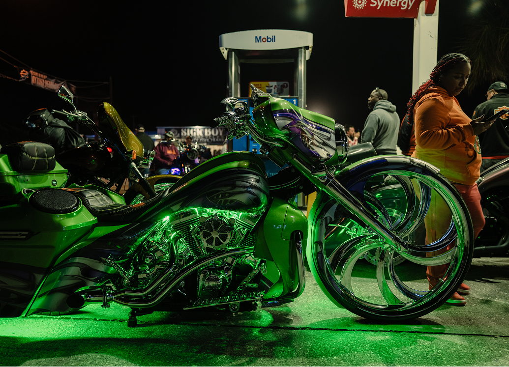 Custom motorcycle illuminated with green accent lighting parked at a busy nighttime gas station event