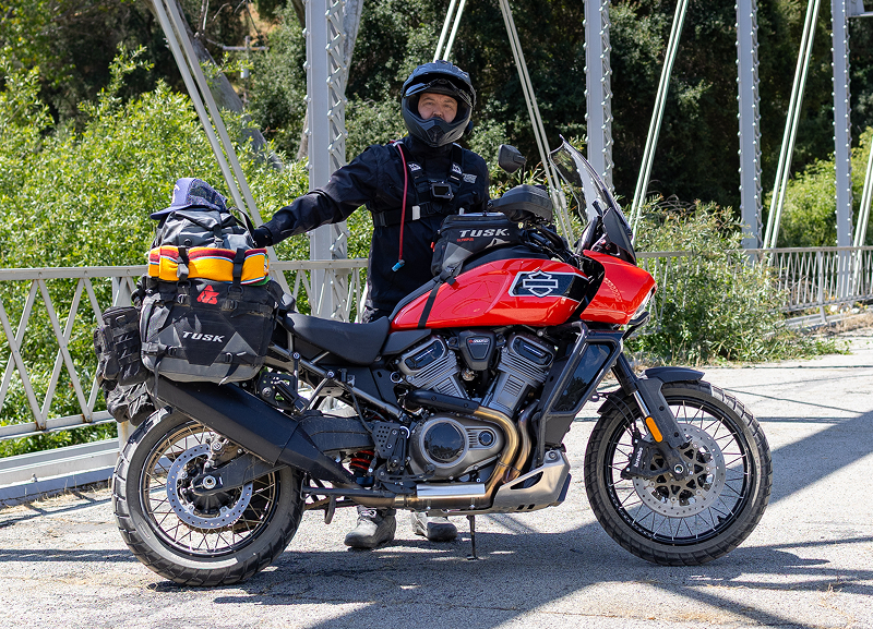 Red H-D Pan America motorcycle parked on a steel bridge with fully loaded luggage, rider standing beside it on sunny day