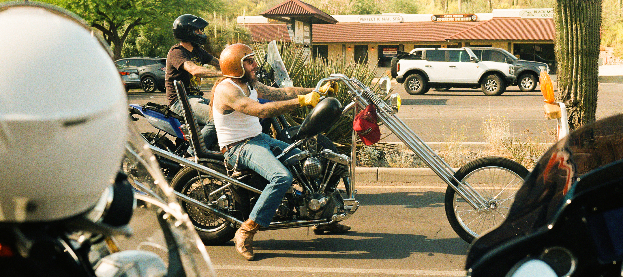 Custom chopper motorcycle ridden through city street, long front fork, helmeted riders, storefronts behind.