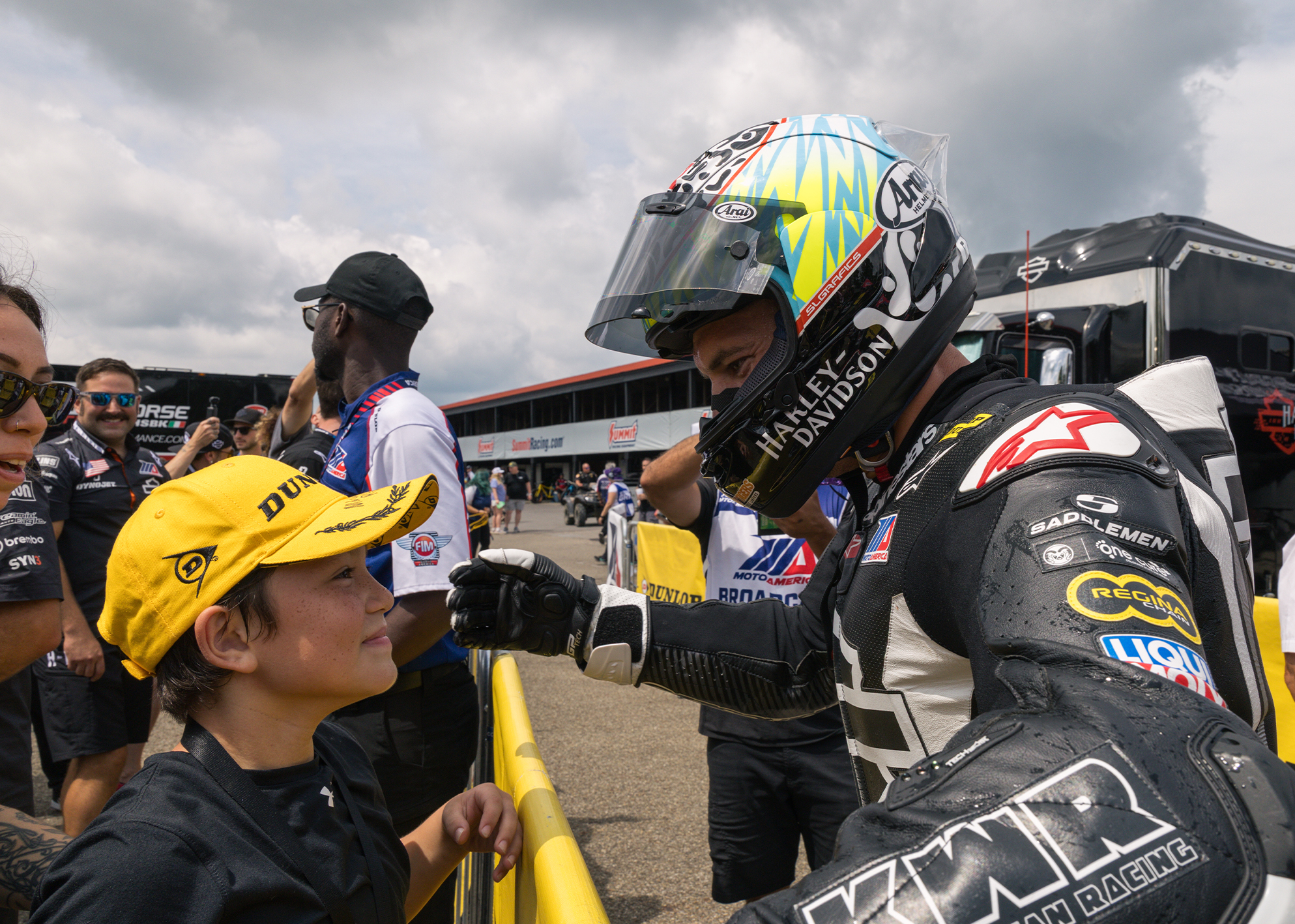 James Rispoli, in full racing gear, shares a fist bump with a young fan wearing a yellow Dunlop cap.