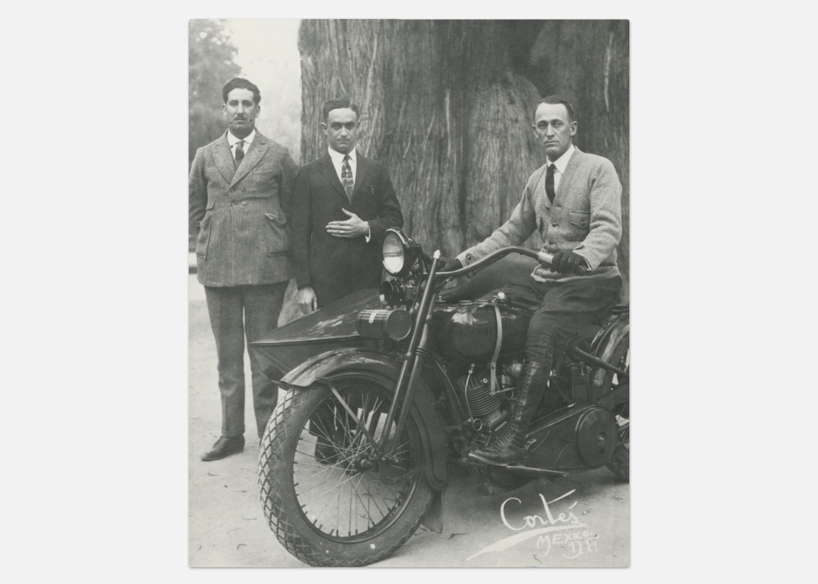 Historic black-and-white photo taken by Mexican photographer Vicente Cortés Sotelo of three men, one seated on a vintage motorcycle beside a massive tree.