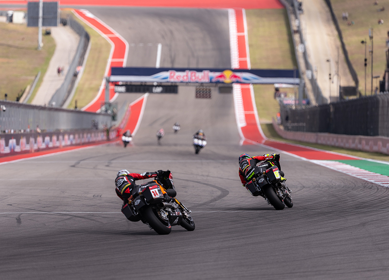 Two motorcycle racers accelerate side by side down a straight away bordered by bright track markings