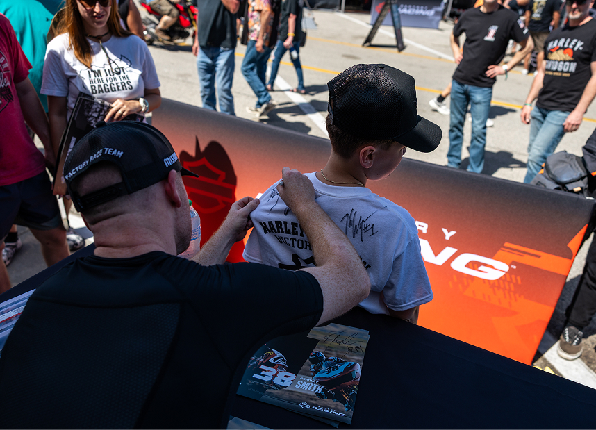 Bradley Smith signs the back of a child’s t-shirt at an event booth, with a crowd gathered behind them
