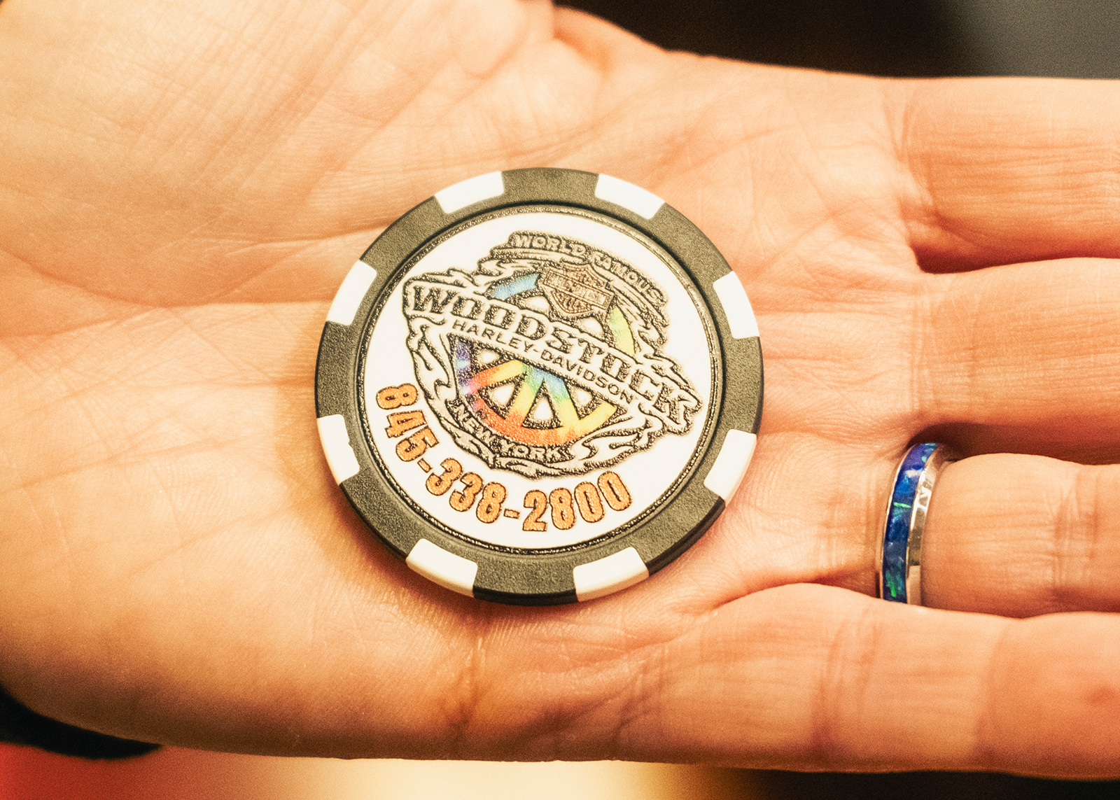 An open hand holds a poker style chip with a Woodstock Harley-Davidson peace logo and phone number.
