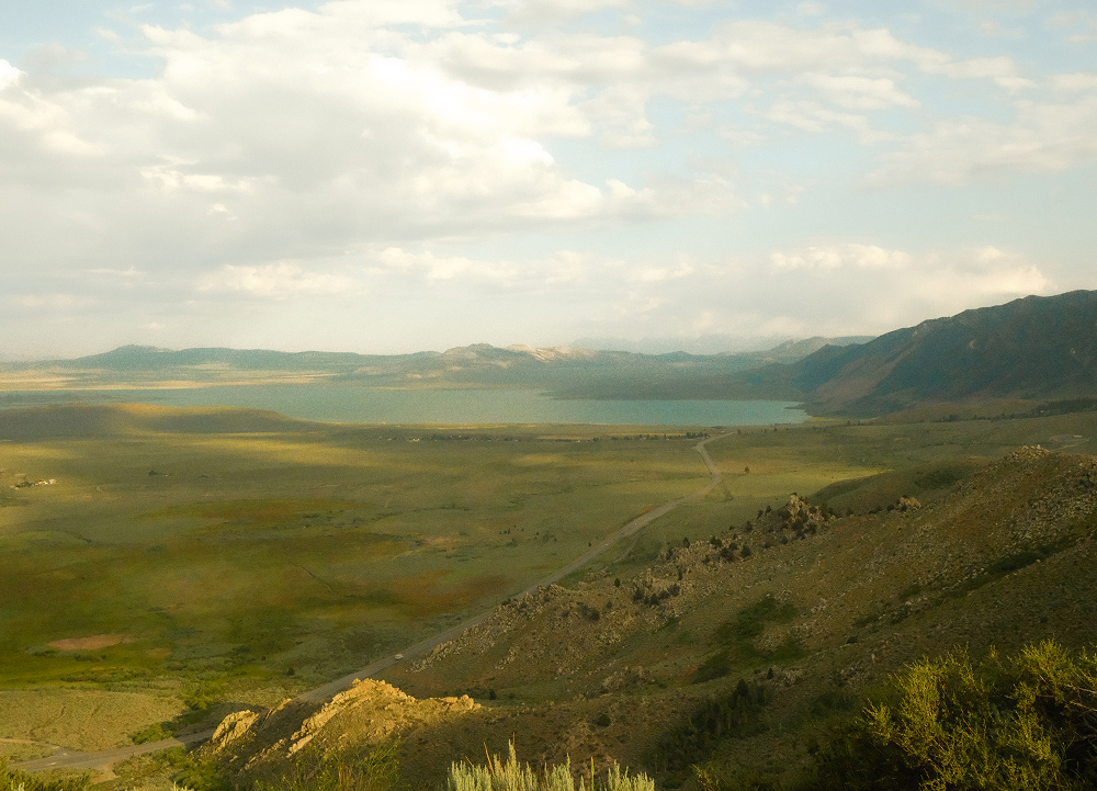 Mountain valley and lake viewed from a hillside overlook, with winding road, open grasslands, and clouds above