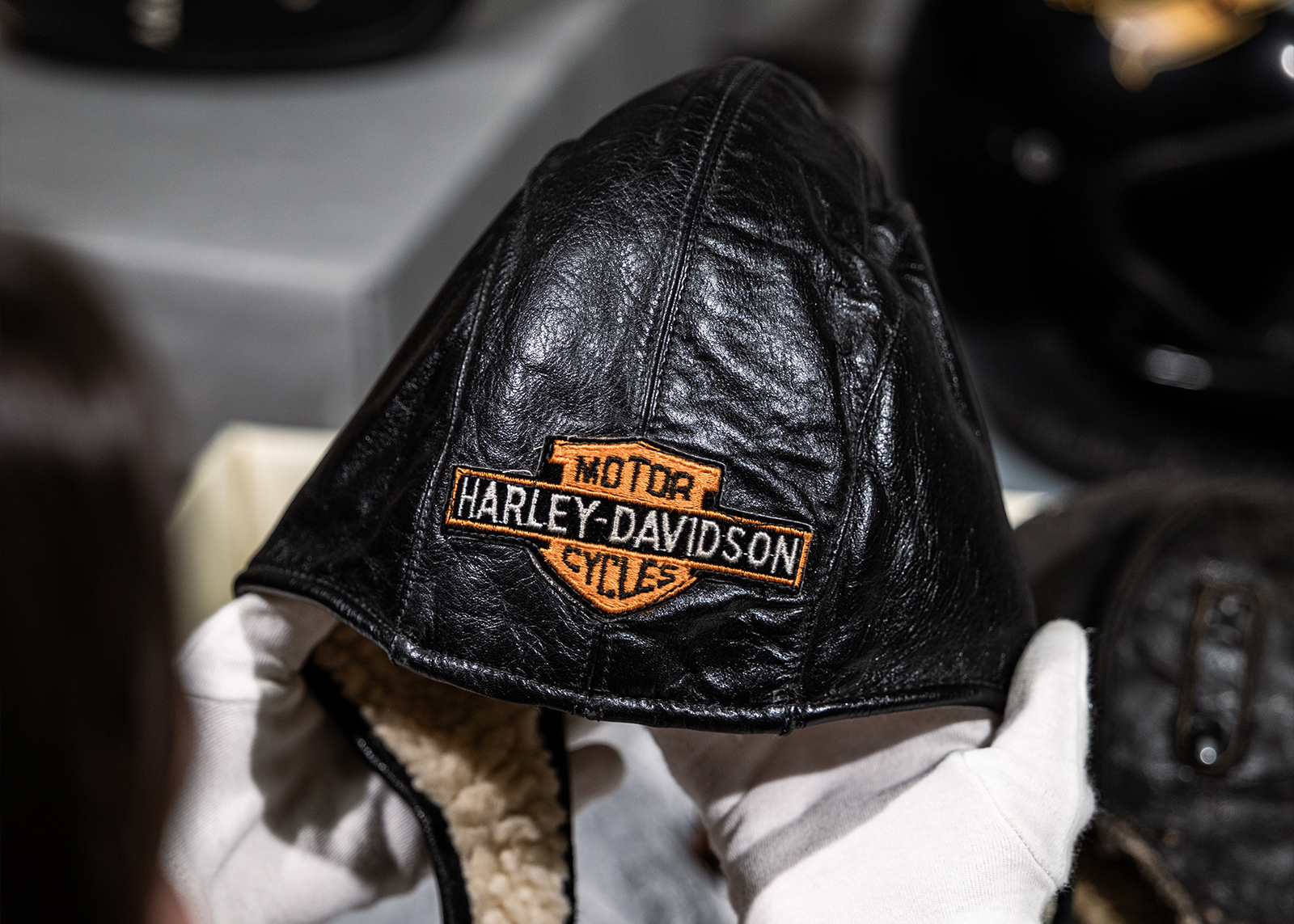 A museum archivist holds a vintage leather motorcycle helmet with Harley-Davidson bar and shield logo embroidered on the front.