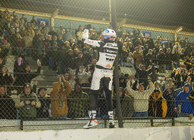 Briar Baumann climbs the catch fence celebrating with raised arm as crowd behind reacts under bright track lighting