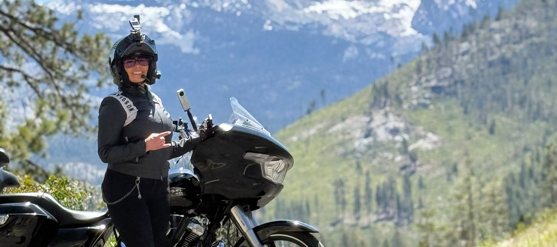 The author stands beside a Harley-Davidson at a mountain overlook with alpine peaks in the distance