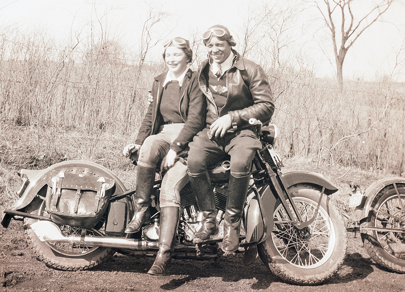 Vintage photo of Gustaf Stenmark and Elsi Toplak seated on a classic motorcycle on a dirt path with sparse trees behind them