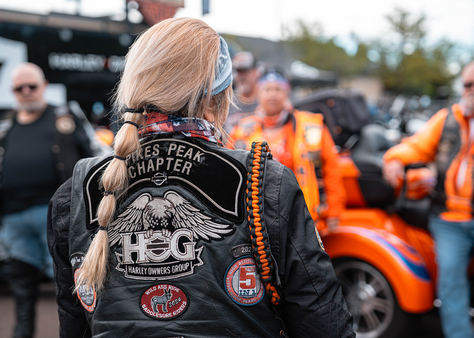 Person with long blonde ponytail wearing a Pikes Peak Chapter H.O.G. jacket stands among motorcycles and other riders