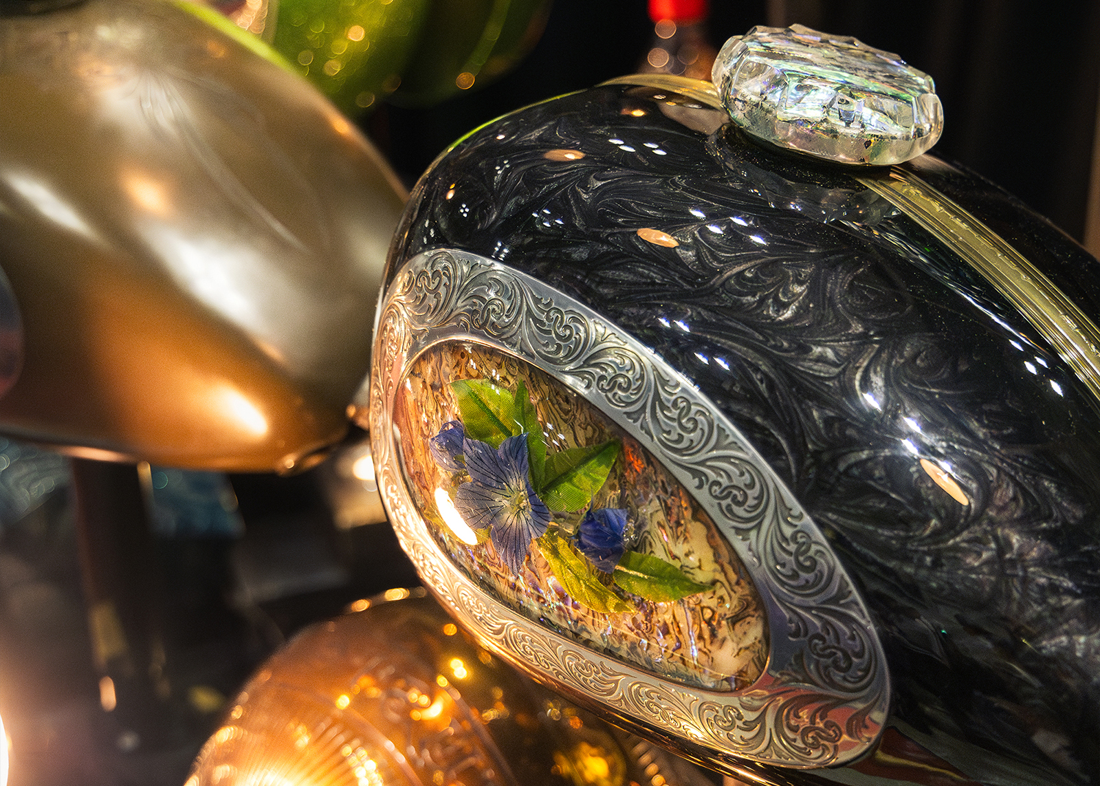 Close-up of a custom motorcycle fuel tank with intricate floral artwork and engraved silver detailing