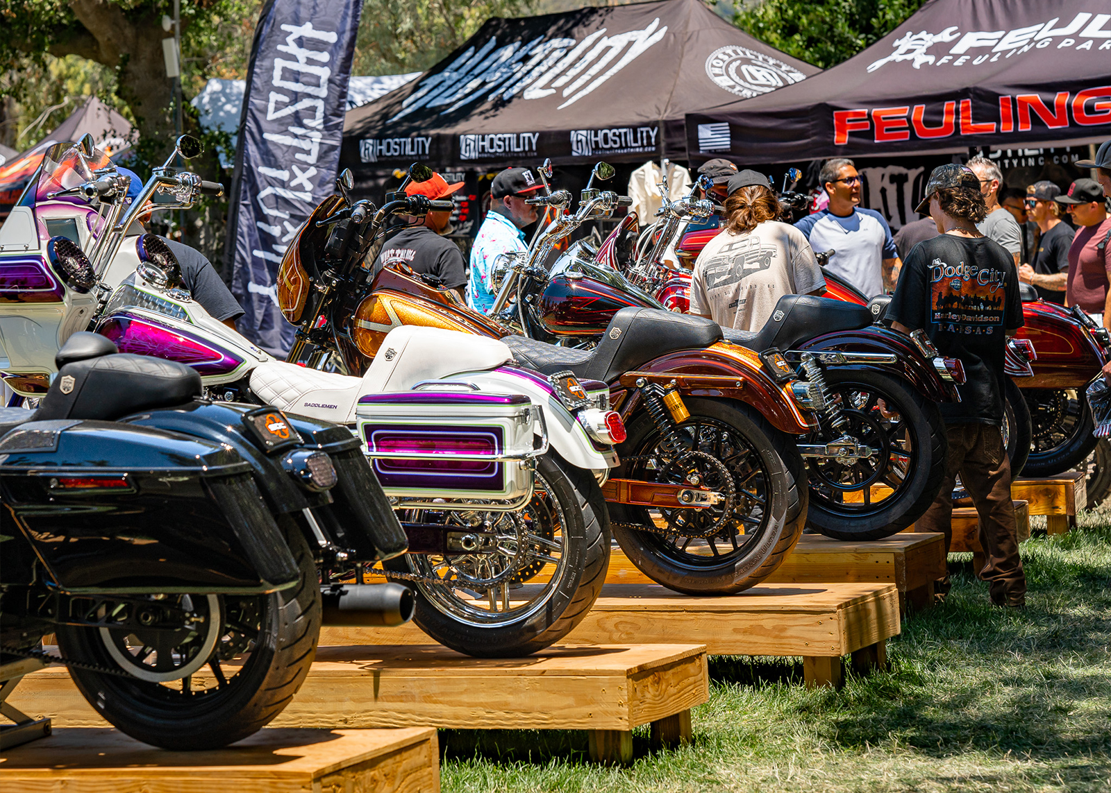 Six colorfully painted Harley’s on wooden stands with onlookers in the background.