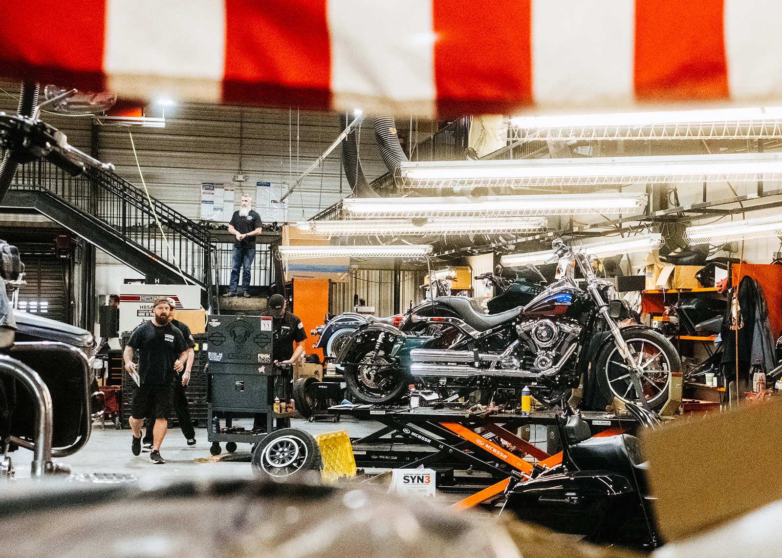 Bikes on lifts and staff in the service department of Moonshine Harley-Davidson, with the edge of an American flag seen hanging in the foreground.