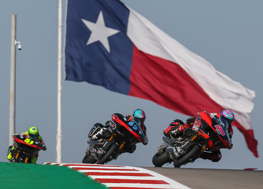 Motorcycle racers lean into a turn on a track, riding beneath a large Texas flag waving overhead