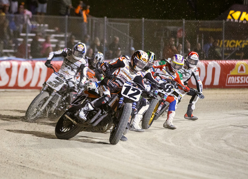 Riders lean deep into a packed dirt corner in tight formation kicking up dust under stadium lights
