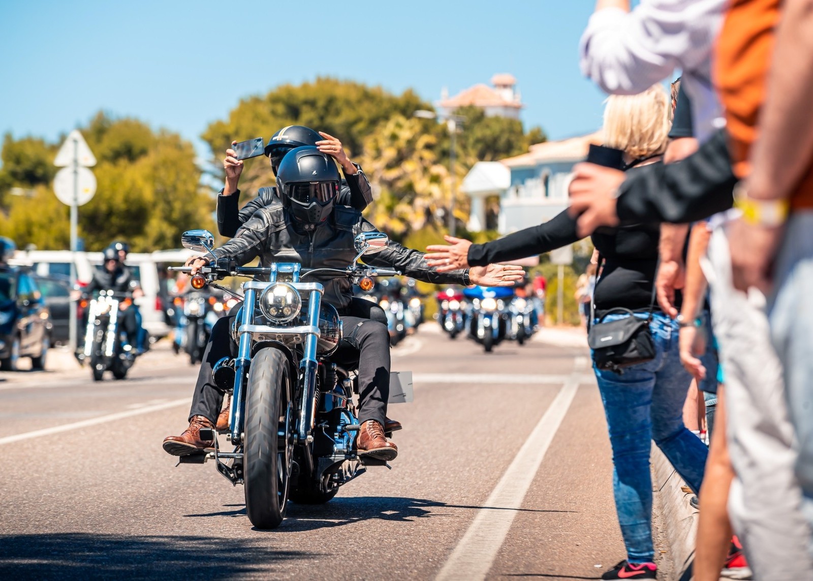 A group of motorcyclists riding down a street while people stand nearby and reach out to greet them.