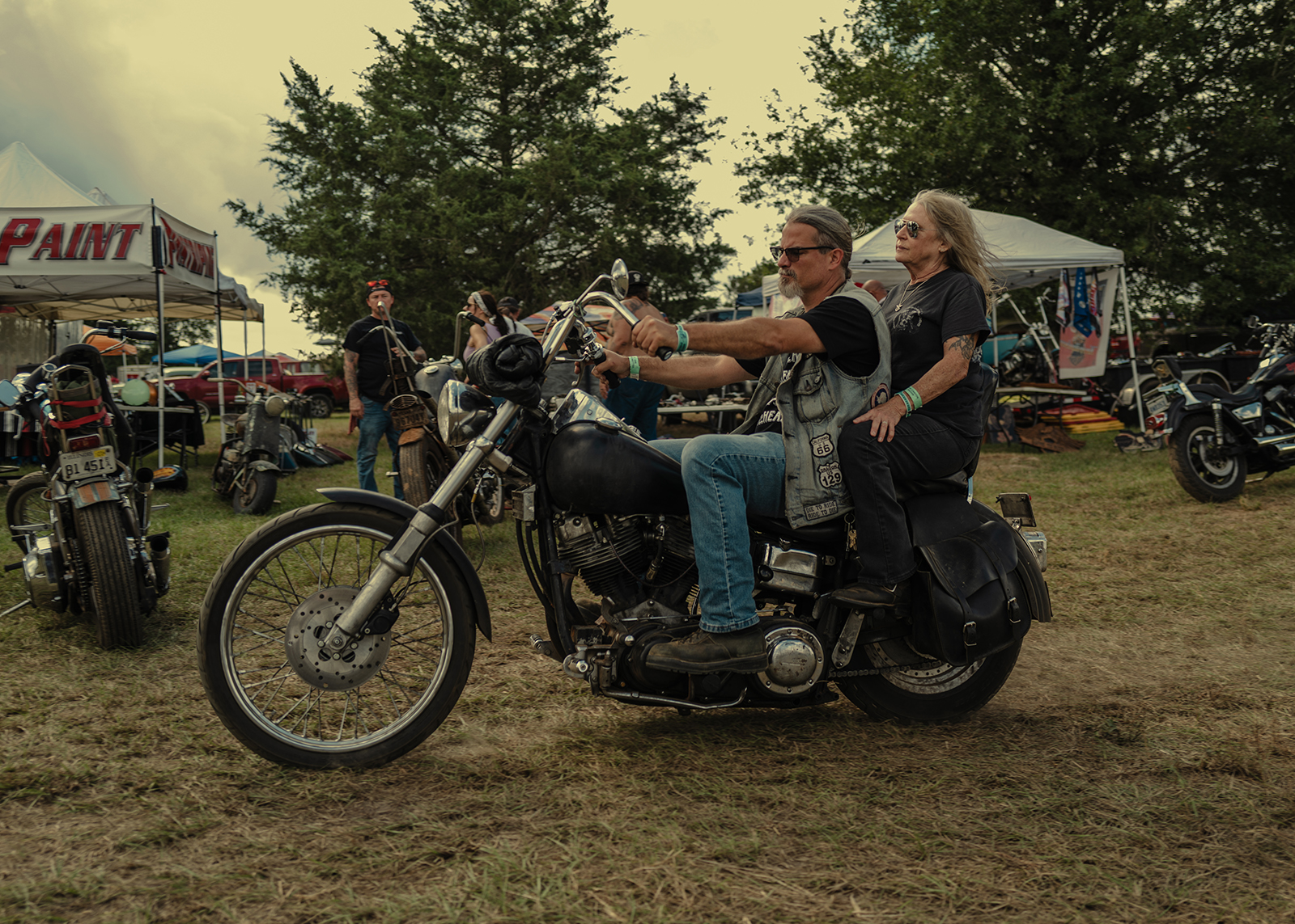 An older couple rides a vintage Harley through a field at the Born Free Texas rally, surrounded by tents, bikes, and fellow riders.