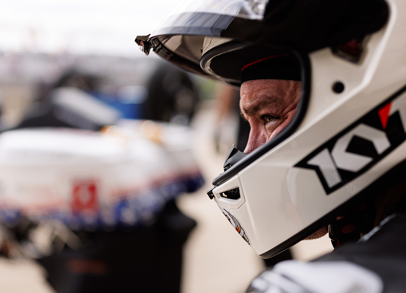 Close-up of a helmeted motorcycle racer in the paddock with team members and equipment blurred behind