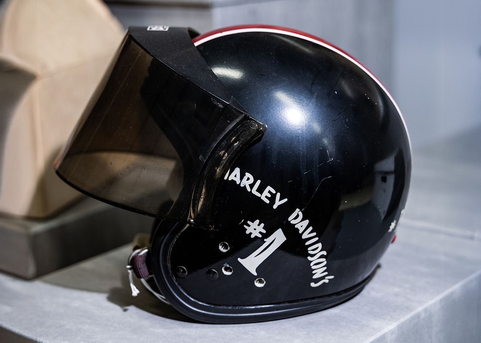 A vintage black plastic motorcycle helmet with a visor, Harley-Davidson #1 hand painted over the ear, and red and white racing stripes going from front to back.