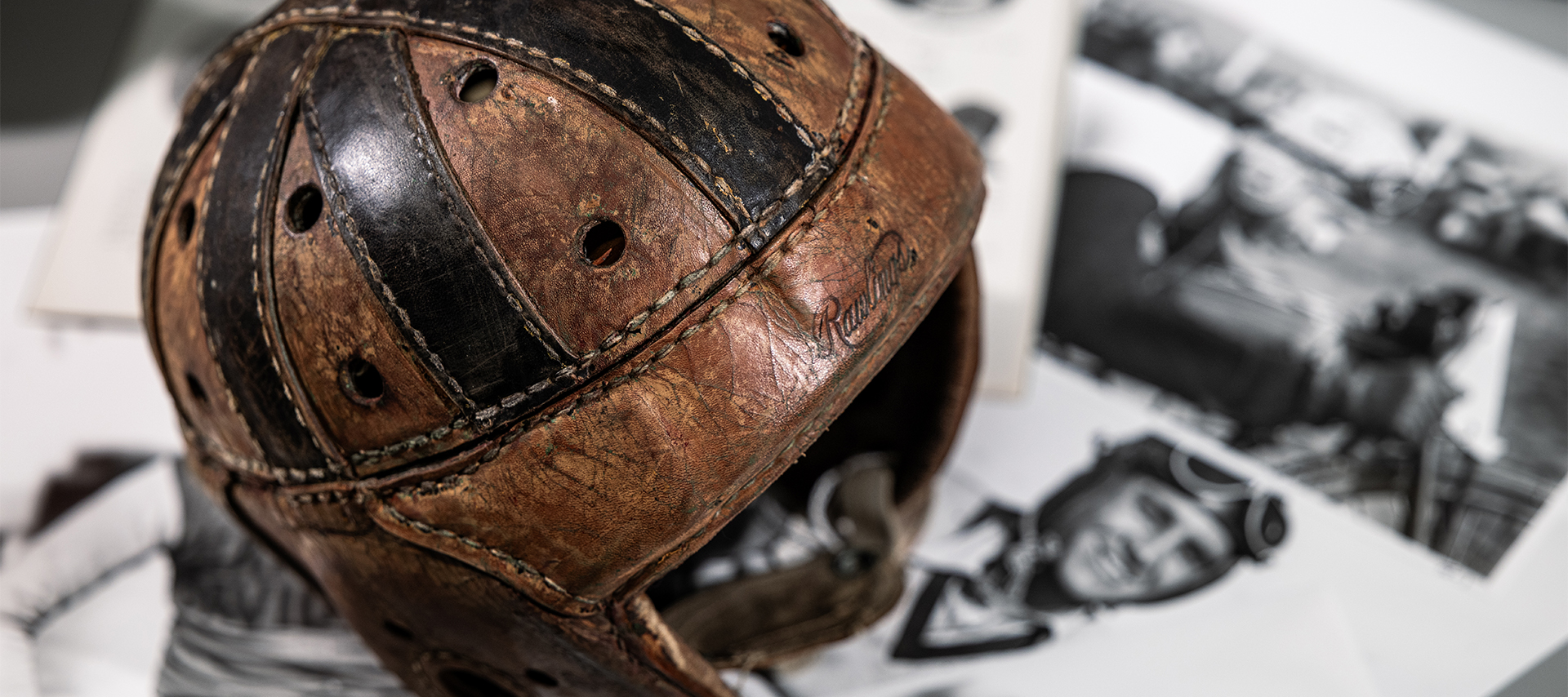 A vintage leather motorcycle helmet with a script Rawlings logo stamped on the front is displayed on a table surrounded by black and white archival photos.