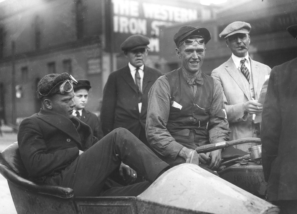 Vintage photo of people in flat caps, vests, jackets, and ties on and around a motorcycle with a sidecar