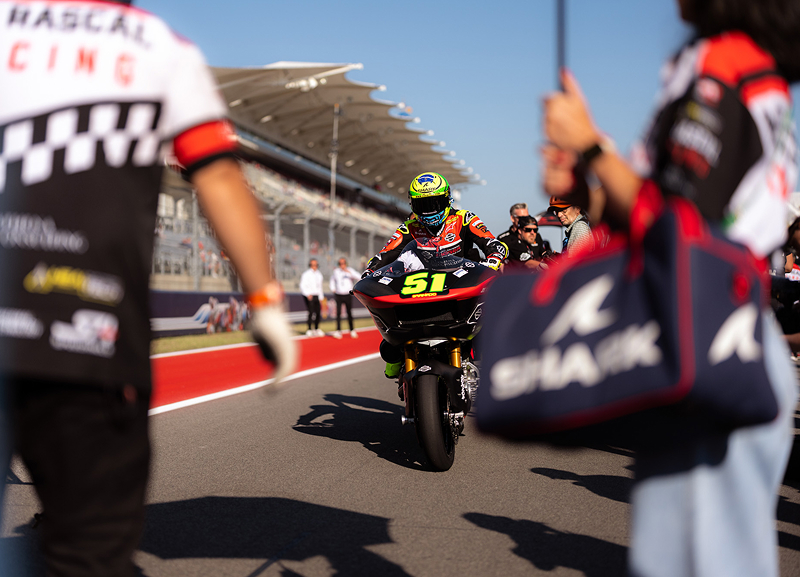Motorcycle racer rides past crew members and pit lane equipment during race day preparations