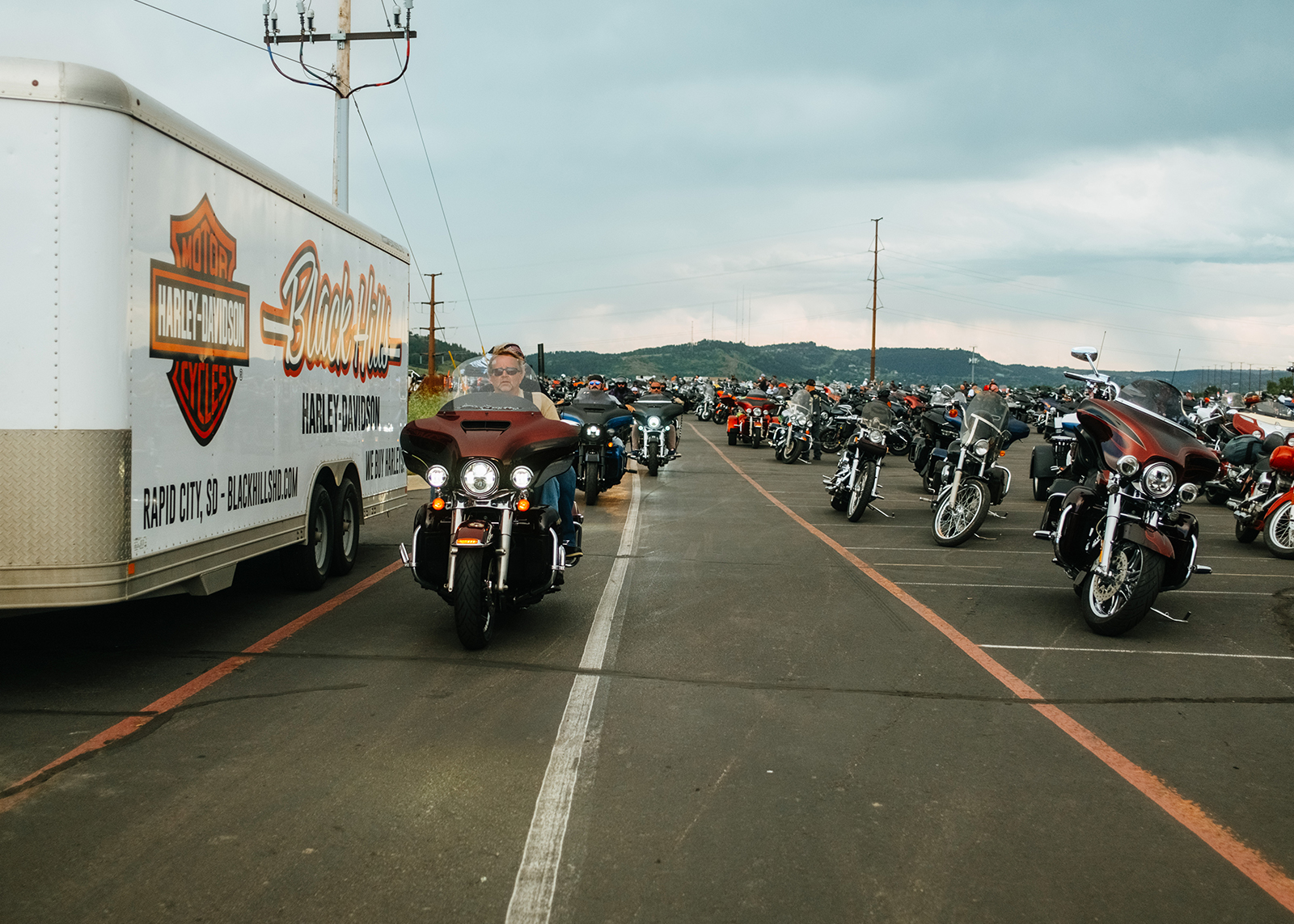 Riders pass a Black Hills Harley-Davidson branded trailer, with rows of motorcycles parked behind.