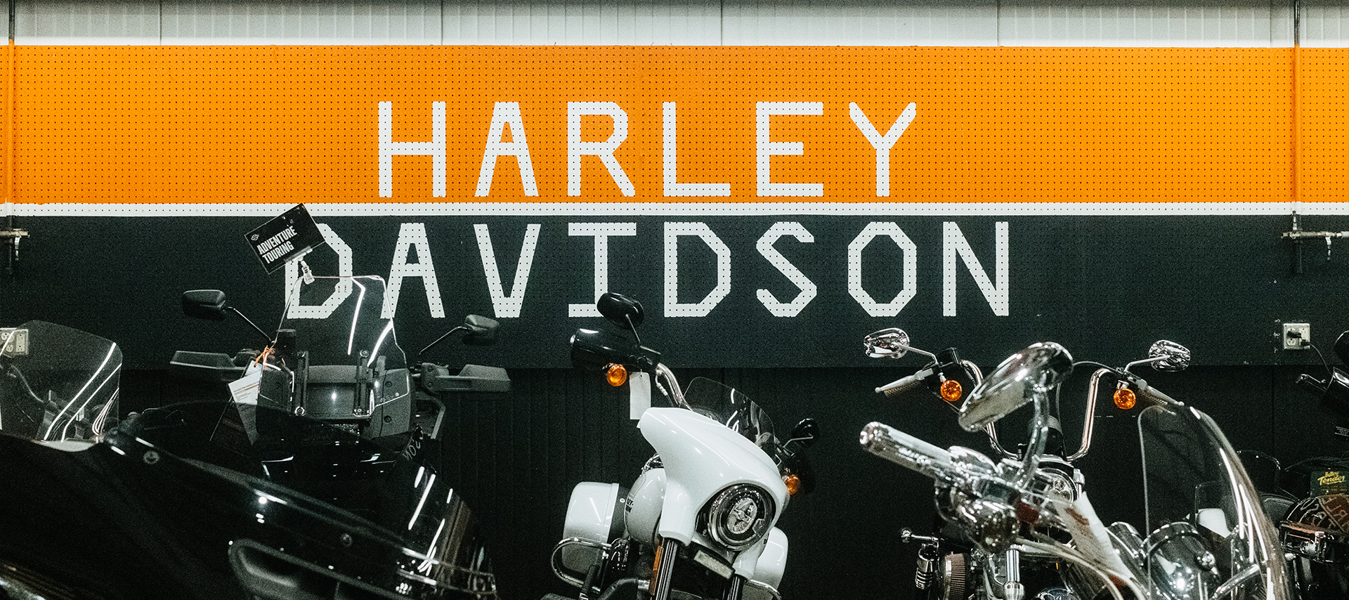 A pegboard painted orange, black, and white that says Harley-Davidson sits behind a row of motorcycles.