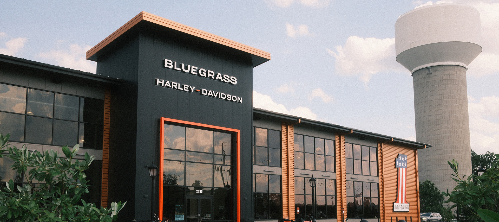 Exterior view of Bluegrass Harley-Davidson dealership with black and orange facade, large windows, and water tower nearby