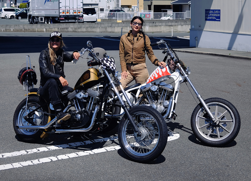 Two custom choppers parked side by side in a parking lot with the authors posing beside them in bright daylight