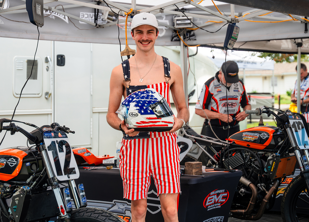 Racer Kody Kopp in patriotic overalls holds a helmet beside flat track motorcycles in the team paddock