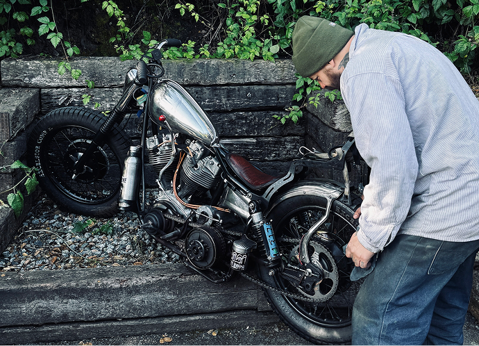 Christian Sosa working on hand-built 1976 Shovelhead beside stacked wooden beams with greenery in a shaded outdoor setting