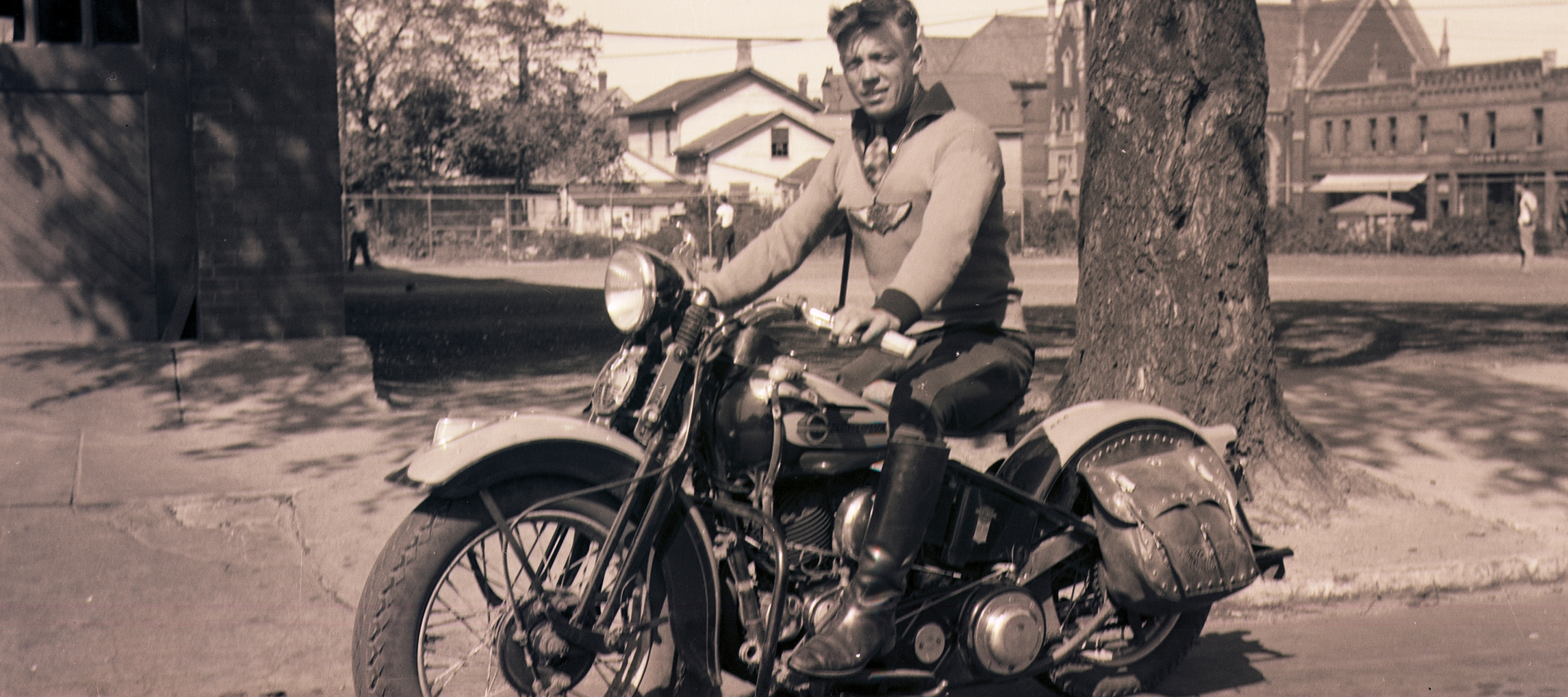 Vintage photo of Gustaf Stenmark seated on a classic motorcycle beside a tree in a small-town street with buildings in the background