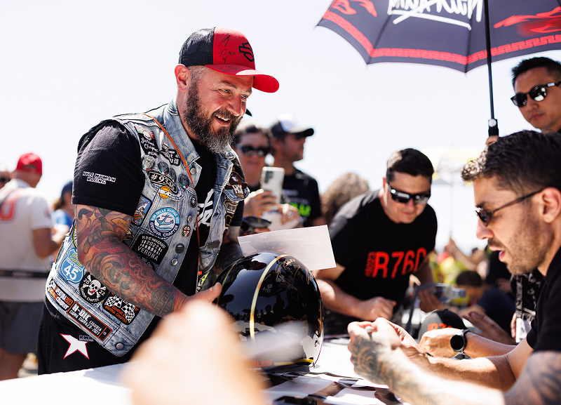 Person in a Harley-Davidson vest gets autographs and chats with a racer during a race weekend event