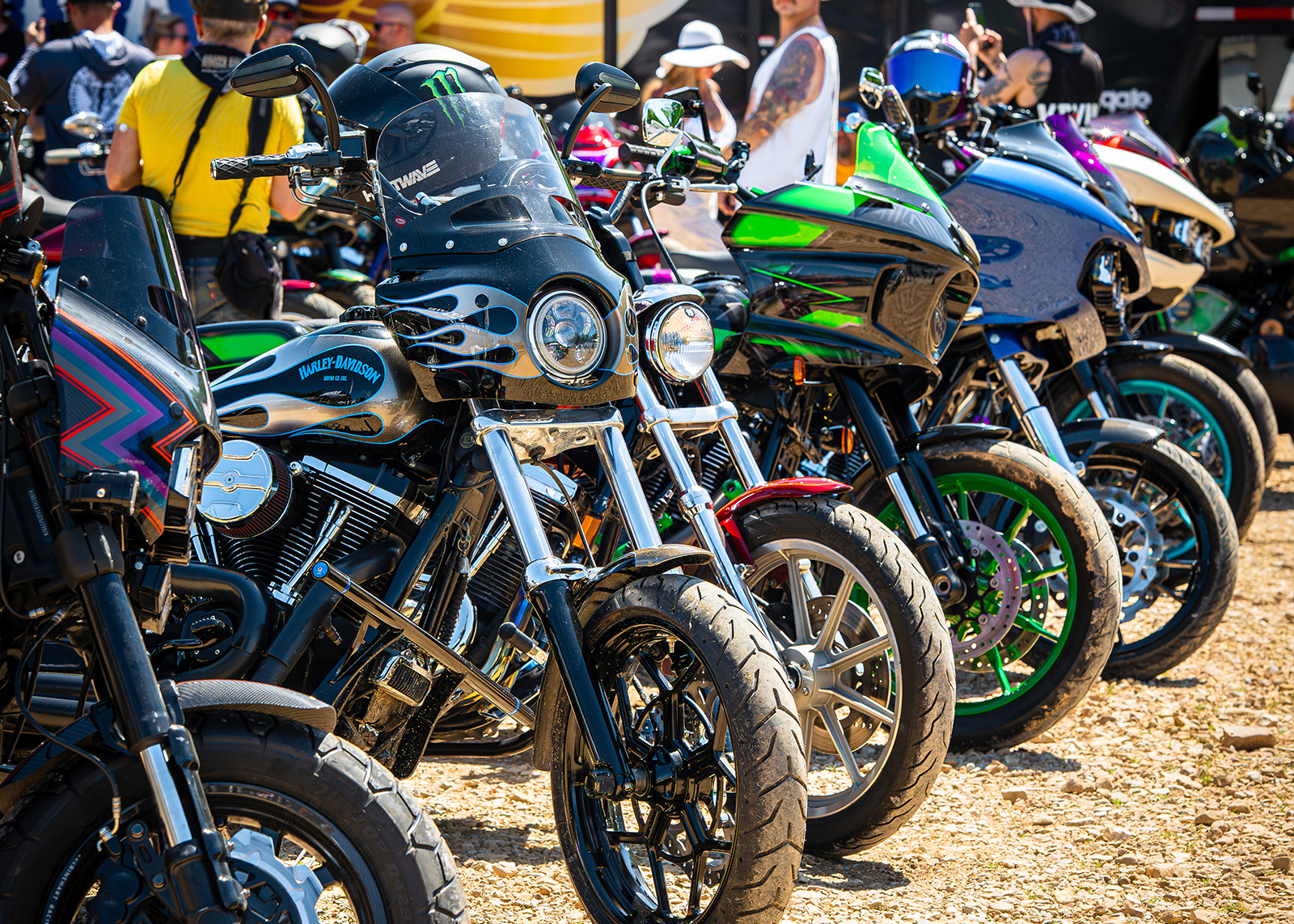 A line of motorcycles parked in the dirt with a crowd behind them.
