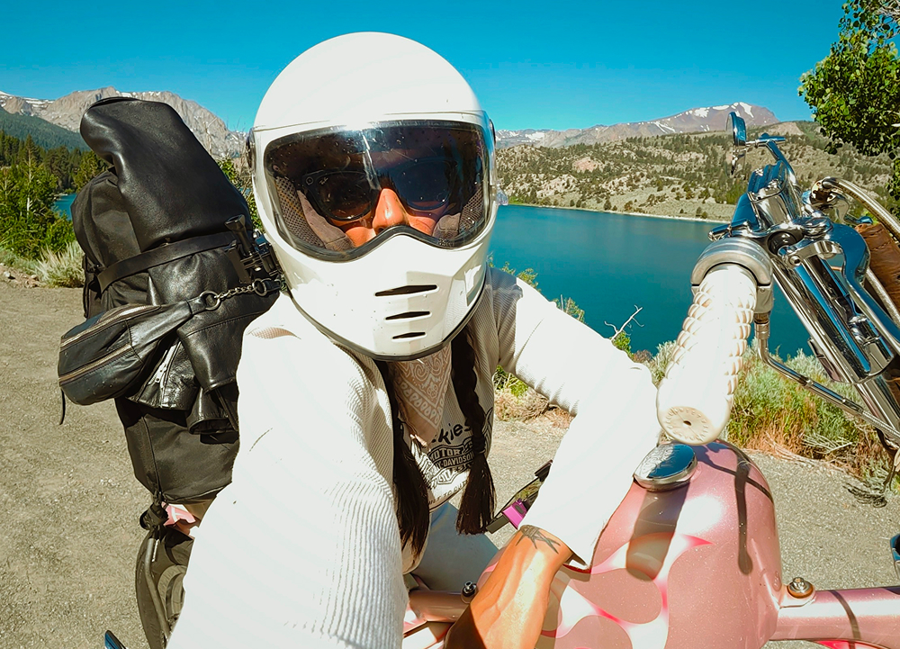 Helmeted rider seated on a custom chopper beside a turquoise alpine lake with forested mountains beyond