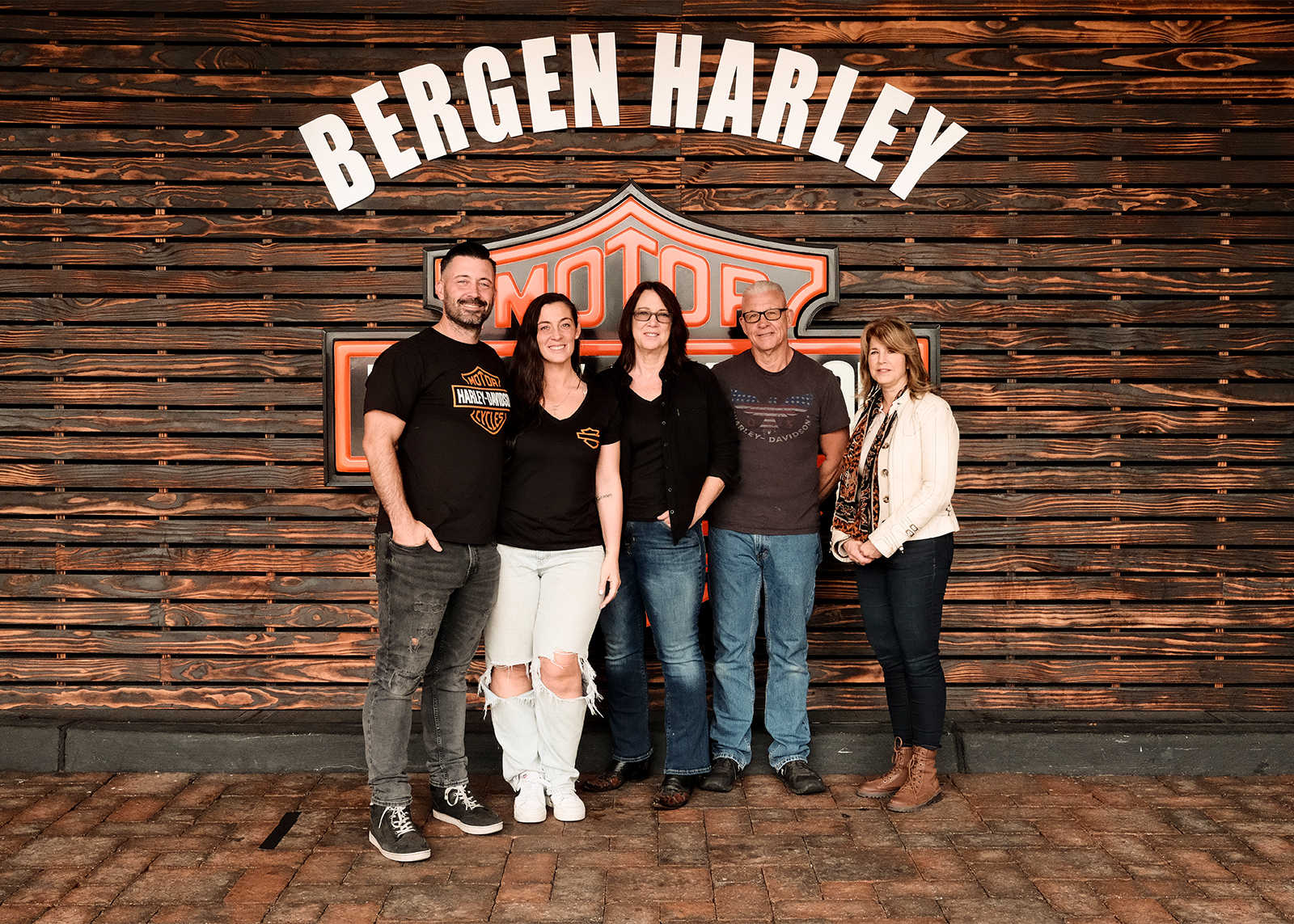5 core members of the Bergen Harley-Davidson family pose in front of a dealership sign on a wood paneled wall.
